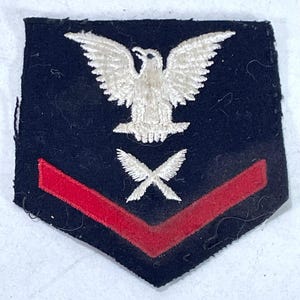 Puede incluir: Parche azul marino con un águila bordada en blanco, alas blancas cruzadas y un galón rojo. Es probable que sea una insignia de rango militar.