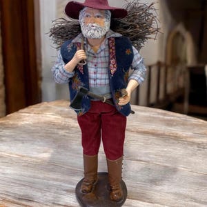 May include: A figurine of an elderly man with a white beard and a burgundy hat, carrying a bundle of twigs on his back. He wears a plaid shirt, a blue vest with floral embroidery, red corduroy pants, and brown boots. An axe hangs from his belt.