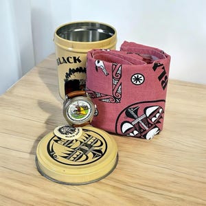 May include: A collection of items including a tan metal container with the word "BLACK" printed on it, a rolled red bandana with a black and white design, a watch with a brown leather band, and a yellow lid with a black graphic.
