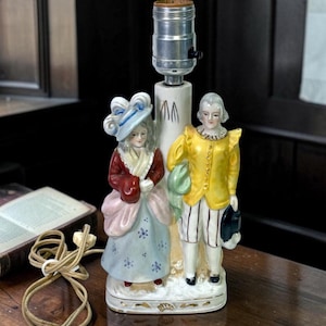 May include: A porcelain table lamp base featuring two figures standing side by side. The left figure wears a blue hat, red jacket, and light blue skirt. The right figure wears a yellow jacket and white striped trousers. A metal socket sits atop.