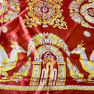 May include: A vibrant red textile with intricate gold and white embroidery. The design features symmetrical patterns, including stylized mythical creatures, floral motifs, and ornate archways. The fabric has a silky sheen and a fringed edge.