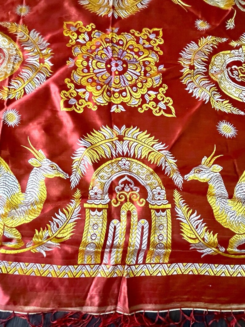 May include: A vibrant red textile with intricate gold and white embroidery. The design features symmetrical patterns, including stylized mythical creatures, floral motifs, and ornate archways. The fabric has a silky sheen and a fringed edge.