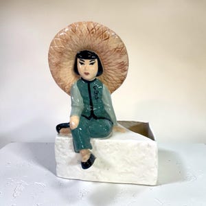May include: A ceramic figurine of a person wearing a green outfit and a large straw hat, seated on a white rectangular planter. The figurine has black hair and is wearing black shoes. The planter has a textured surface.