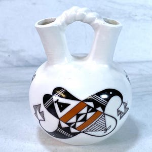May include: White ceramic vase with two spouts and a handle. The vase features a black, white, and orange geometric design. The vase is a decorative piece, likely for display or holding flowers.