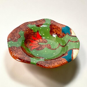 May include: A small, shallow ceramic bowl with an irregular, wavy rim. The interior features a vibrant green base with small yellow dots, decorated with large, textured brown patches, bright red floral accents, and solid teal blue shapes.