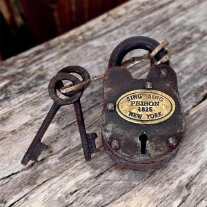 Op de afbeelding: Een antiek, verweerd hangslot met twee sleutels. Het donkerbruine slot heeft een messing plaatje met de tekst "Sing Sing Prison 1825 New York". De sleutels zijn bevestigd en vertonen tekenen van ouderdom, wat wijst op een vintage item.