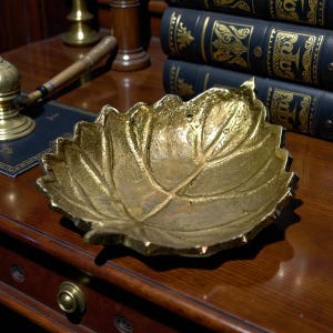 May include: A gold-colored leaf-shaped decorative bowl. The bowl has a textured surface and is resting on a wooden surface. The bowl is likely made of metal and is a decorative item.