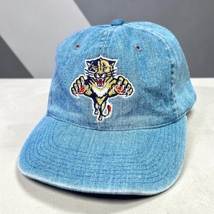May include: A light blue denim baseball cap featuring an embroidered graphic of a leaping panther with gold, white, and red details. The cap has a curved brim, a button on the top, and stitched eyelets for ventilation, showcasing a classic six-panel design.
