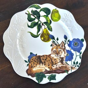 Op de afbeelding: Witte decoratieve plaat met een geschulpte rand. De plaat heeft een handgeschilderd ontwerp van een bobcat, peren en blauwe bloemen. De bobcat is bruin met zwarte vlekken en ligt op een boomstam. De peren zijn groen en geel.