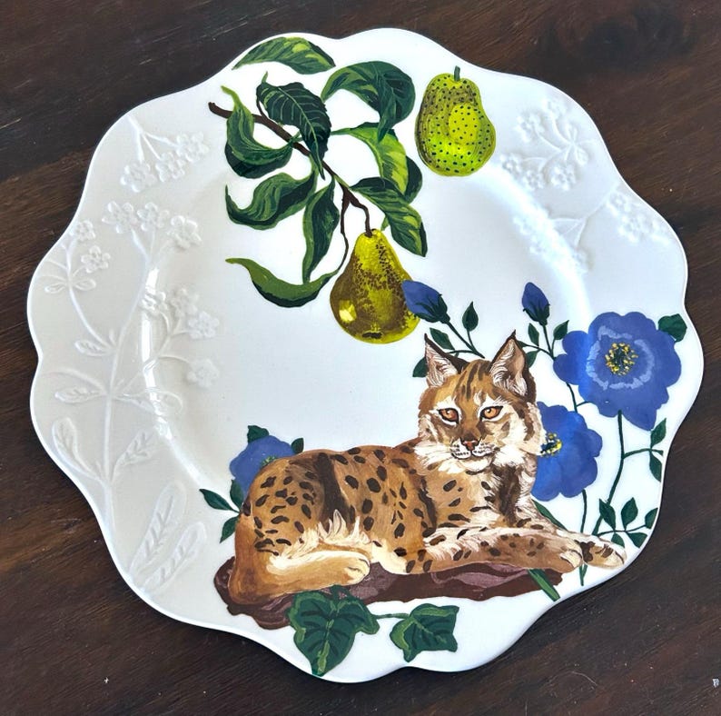 Op de afbeelding: Witte decoratieve plaat met een geschulpte rand. De plaat heeft een handgeschilderd ontwerp van een bobcat, peren en blauwe bloemen. De bobcat is bruin met zwarte vlekken en ligt op een boomstam. De peren zijn groen en geel.