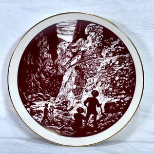 May include: Decorative plate with a maroon and white illustration of a train traveling through a mountain pass. The plate has a gold rim and features figures of people in the foreground.