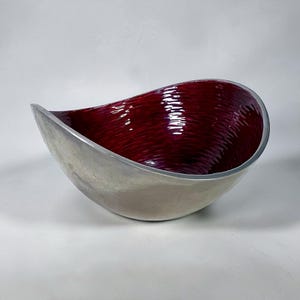 May include: A modern, teardrop-shaped bowl with a silver exterior and a deep red interior. The interior has a textured, rippled surface. The bowl is made of metal and is suitable for decorative use.