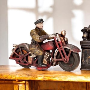 May include: A vintage cast iron motorcycle toy with a rider. The motorcycle is primarily red with black tires and silver accents. The rider is wearing a khaki uniform and a black cap. The toy is sitting on a wooden surface.