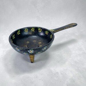Op de afbeelding: Een kleine, zwarte gietijzeren pan met een lange steel en drie korte pootjes. De pan is versierd met handgeschilderde gele, blauwe en witte bloemmotieven rond de rand. De steel heeft gouden accenten.