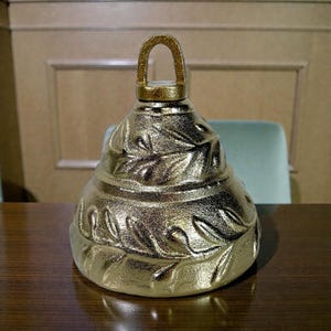 Op de afbeelding: Een goudkleurige decoratieve bel met een lusvormige handgreep. De bel heeft een textuur met een bladerenpatroon. De bel staat op een bruin houten oppervlak.