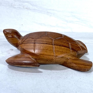 May include: A handcrafted wooden turtle figurine. The turtle is carved from a light brown wood, with visible grain and a detailed shell pattern. The figurine has a smooth finish and is posed with its head and flippers extended.
