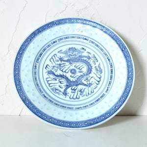 May include: A round ceramic plate featuring a central blue and white illustration of a dragon surrounded by clouds. The rim is decorated with a geometric cross-hatch pattern and a series of small, translucent circular cutouts along the edge.