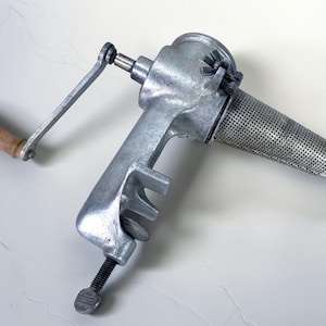May include: A vintage, silver-colored food mill with a wooden handle and a perforated cone-shaped strainer. The mill has a clamp for securing it to a counter and a crank for turning the mechanism. It is a kitchen tool.