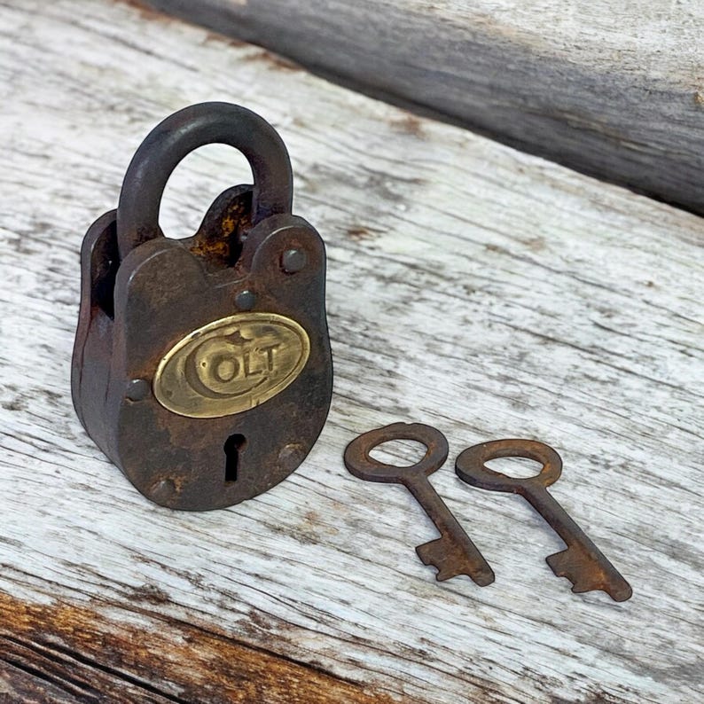 Op de afbeelding: Een vintage, roestig hangslot met een messing plaatje met de tekst "OLT" en twee bijpassende sleutels. Het slot is donkerbruin met een gebogen beugel. De sleutels zijn ook roestbruin.