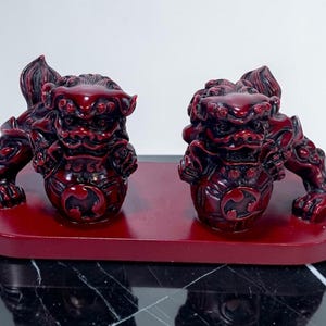 May include: Two red Foo Dog statues on a matching red base. The statues are detailed with intricate carvings and a glossy finish. The Foo Dogs are positioned facing forward, with their paws resting on the base.
