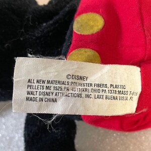Puede incluir: Primer plano de un peluche de Disney. El juguete tiene tela negra, roja y blanca. La tela roja tiene c&iacute;rculos amarillos. Una etiqueta blanca con el logotipo de Disney y texto sobre materiales y origen es visible.