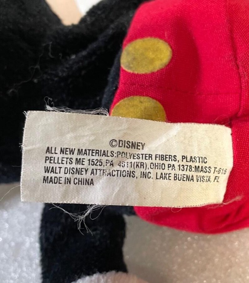 Puede incluir: Primer plano de un peluche de Disney. El juguete tiene tela negra, roja y blanca. La tela roja tiene c&iacute;rculos amarillos. Una etiqueta blanca con el logotipo de Disney y texto sobre materiales y origen es visible.