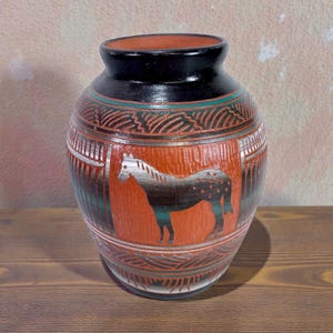 Op de afbeelding: Een handgemaakte keramische vaas met een paardenontwerp. De vaas heeft een zwarte rand, een rode body met een paardensilhouet en groene en zilveren accenten. De vaas is een decoratief item.