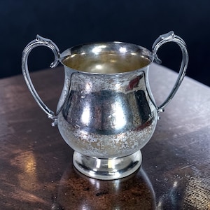 May include: A silver-colored sugar bowl with two curved handles. The bowl has a rounded body and sits on a small, circular base. The interior of the bowl is a shiny silver color, and the exterior shows signs of wear. The handles have a decorative, curved design.