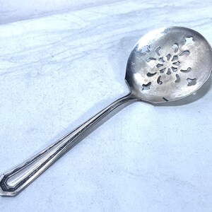 May include: A silver-colored serving spoon with a snowflake-shaped design in the bowl. The handle has a decorative, geometric design. The spoon is likely made of stainless steel or a similar metal.