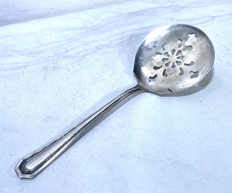 May include: A silver-colored serving spoon with a snowflake-shaped design in the bowl. The handle has a decorative, geometric design. The spoon is likely made of stainless steel or a similar metal.