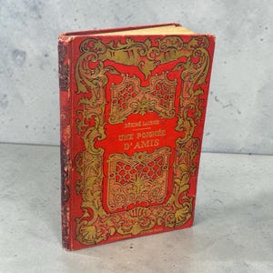May include: A red hardcover book titled Une Poignée d'Amis by Désiré Lacroix. The front cover features intricate gold embossed scrollwork and decorative patterns framing the central text. The spine shows signs of wear with dark red detailing.
