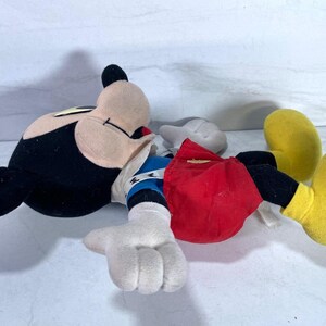 May include: A plush Mickey Mouse doll, lying on its back. The doll features a black head and ears, a tan face, white gloves, red shorts, and yellow shoes. The doll's arms are outstretched, and it appears to be in a relaxed position.