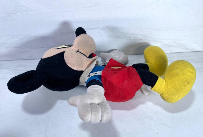 May include: A plush Mickey Mouse doll, lying on its back. The doll features a black head and ears, a tan face, white gloves, red shorts, and yellow shoes. The doll's arms are outstretched, and it appears to be in a relaxed position.