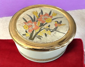 Könnte beinhalten: Eine runde, weiße Schmuckdose mit goldenem Rand und Deckel. Der Deckel zeigt ein Blumendesign mit orangefarbenen, gelben und grünen Blumen und einem kleinen Vogel. Die Dose steht auf einer roten Oberfläche.