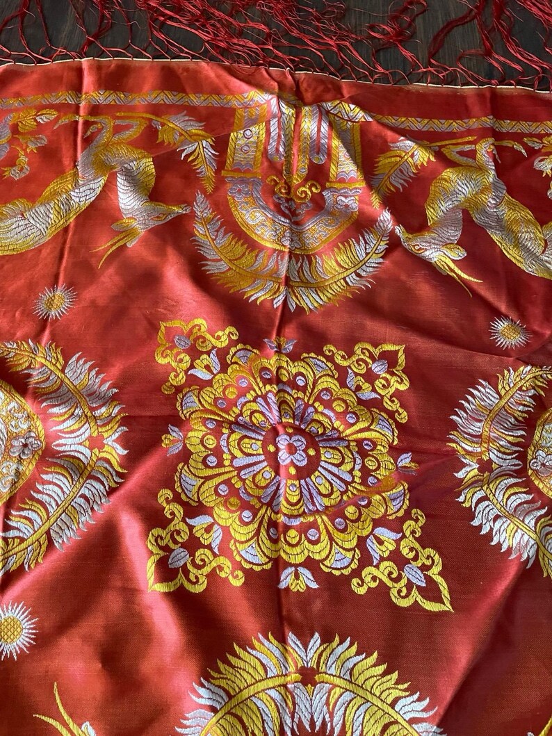 May include: A red textile with intricate gold and silver embroidery. The design features stylized birds, floral patterns, and sunburst motifs. The top edge has a fringe of red threads. The fabric has a lustrous sheen.