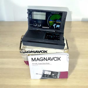 Può includere: Una radiosveglia da viaggio Magnavox AE 4230 nera con display digitale e selettore di fuso orario. La radiosveglia è aperta, rivelando lo schermo e i comandi. La scatola è visibile, con il logo Magnavox e le informazioni sul prodotto.
