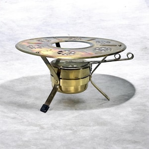 Könnte beinhalten: Ein Vintage-Chafing-Dish aus Messing mit einer runden, perforierten Oberseite und einem Brennstoffbrenner darunter. Das Gericht hat einen dreibeinigen Ständer und zwei Griffe. Das Messing hat einen warmen, goldenen Farbton, der auf Alter hindeutet. Funktionales und dekoratives Design.