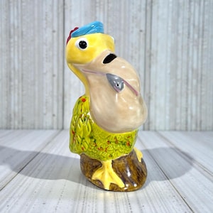 Könnte beinhalten: Eine Keramik-Vogelfigur mit gelbem Kopf, beigem Schnabel und blauem Hut. Der Körper des Vogels ist grün mit roten Akzenten und steht auf einem braunen Sockel mit gelben Füßen. Die Figur hat ein skurriles, cartoonartiges Design.