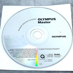 Peut inclure: Un CD de logiciel Olympus Master argenté avec du texte noir indiquant Olympus Master, Olympus et Windows, Macintosh Compatible. Le disque inclut les logos QuickTime, Compact Disc Digital Data et Kodak Online Printing Technology.