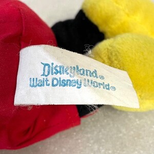 Puede incluir: Primer plano de un peluche con elementos rojos, negros y amarillos. Una etiqueta blanca con el texto "Disneyland, Walt Disney World" es visible. El dise&ntilde;o del juguete sugiere un personaje de Disney.