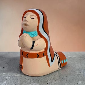 May include: A ceramic figurine of a person in a praying pose. The figure has closed eyes and clasped hands. The figure is painted with orange, white, and turquoise accents. The figure is kneeling.