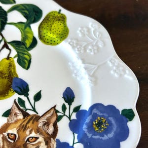 Op de afbeelding: Witte keramische plaat met een geschulpte rand, met een geschilderd ontwerp van een bobcat, peren en blauwe bloemen. De peren zijn groen met zwarte stippen en de bloemen hebben gele centra. De plaat heeft een gedetailleerd, artistiek ontwerp.