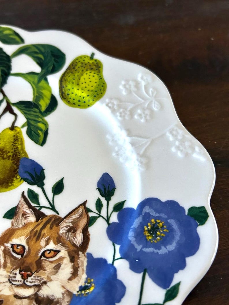 Op de afbeelding: Witte keramische plaat met een geschulpte rand, met een geschilderd ontwerp van een bobcat, peren en blauwe bloemen. De peren zijn groen met zwarte stippen en de bloemen hebben gele centra. De plaat heeft een gedetailleerd, artistiek ontwerp.