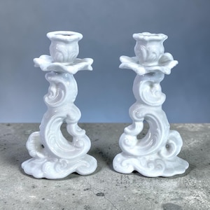 Könnte beinhalten: Zwei weiße Kerzenhalter aus Keramik mit kunstvollen, wirbelnden Designs. Jeder Halter hat eine geriffelte Oberseite und eine Basis mit dekorativen Schnörkeln. Die Kerzenhalter stehen auf einer grauen Oberfläche.