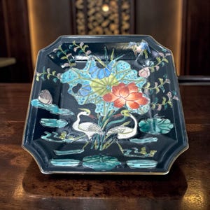 May include: A black, octagonal ceramic dish with a hand-painted floral and bird design. The design features a large red lotus flower, blue and yellow leaves, and two white birds. The dish has a gold trim and is likely used for decorative purposes.