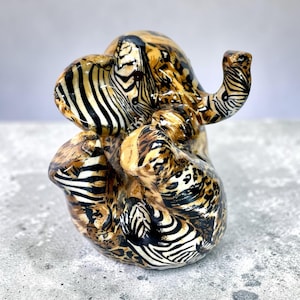 May include: A ceramic elephant figurine with a unique zebra and leopard print design. The sculpture features a glossy finish with black, brown, and tan patterns. The elephant is posed with its trunk raised, showcasing the intricate details of the animal.