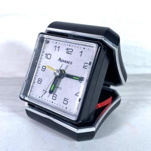 May include: A black and silver travel alarm clock with a white face. The clock face displays the time with black numerals and hands. The word "ADVANCE" is printed on the clock face. The clock is in an open position.