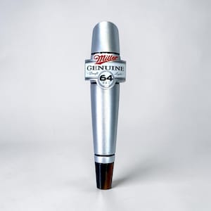 May include: A silver Miller Genuine Draft 64 beer tap handle. The handle has a cylindrical design with a black and brown base. The text "Miller Genuine Draft 64" is displayed on a white and black label with red accents.