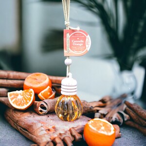 May include: A car air freshener with a cinnamon orange scent. The diffuser is a small glass bottle with a white cap and a string for hanging. The bottle is surrounded by cinnamon sticks and orange slices.