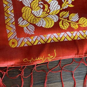 May include: Close-up of a red textile with intricate floral embroidery in yellow and white. The design includes a large flower and geometric patterns. Red fringe hangs from the bottom edge. The textile has text in gold.